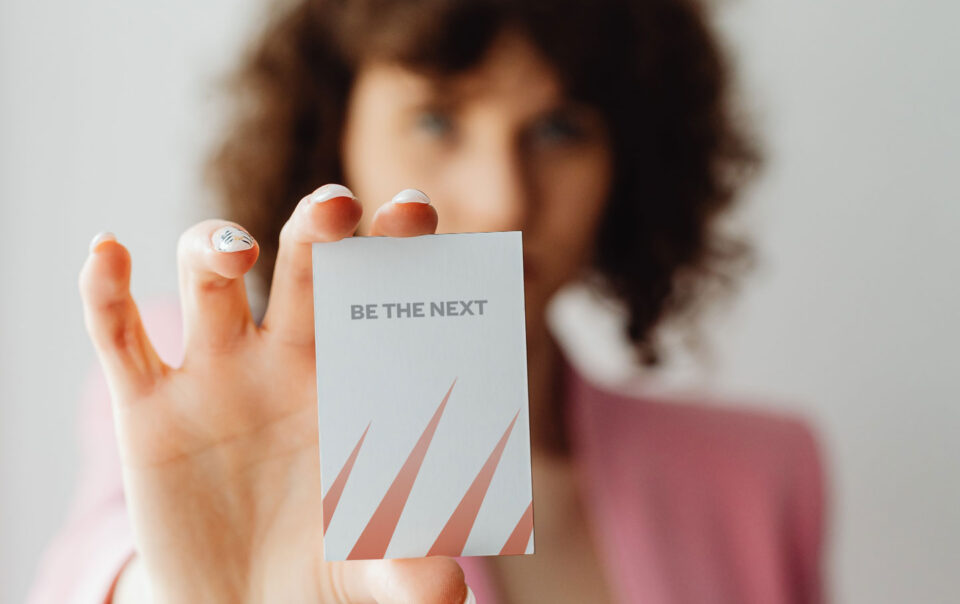 Woman holding a business card with the words "BE THE NEXT" on it.