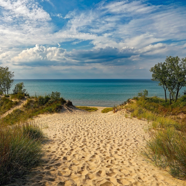 A sandy beach path to water with a blue sky and clouds.