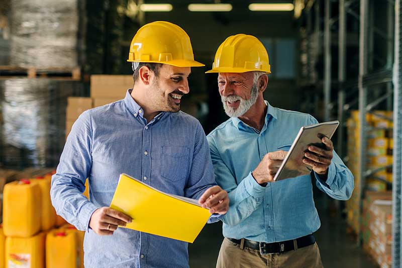 Two successful smiling business man walking through big warehouse with helmets on their heads. Older man is holding digital tablet and shoving younger one some documents.