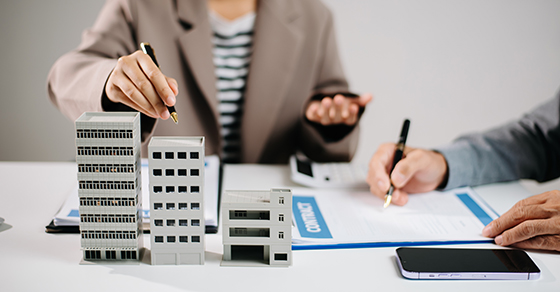 Two individuals at a table. One is writing on a notepad while the other is pointing at two model commercial office buildings.