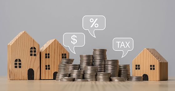 Wooden model houses surround stacks of coins on a tabletop, with speech bubbles displaying a dollar sign, percentage symbol, and the word "TAX", illustrating real estate investing, taxes, and financial planning.
